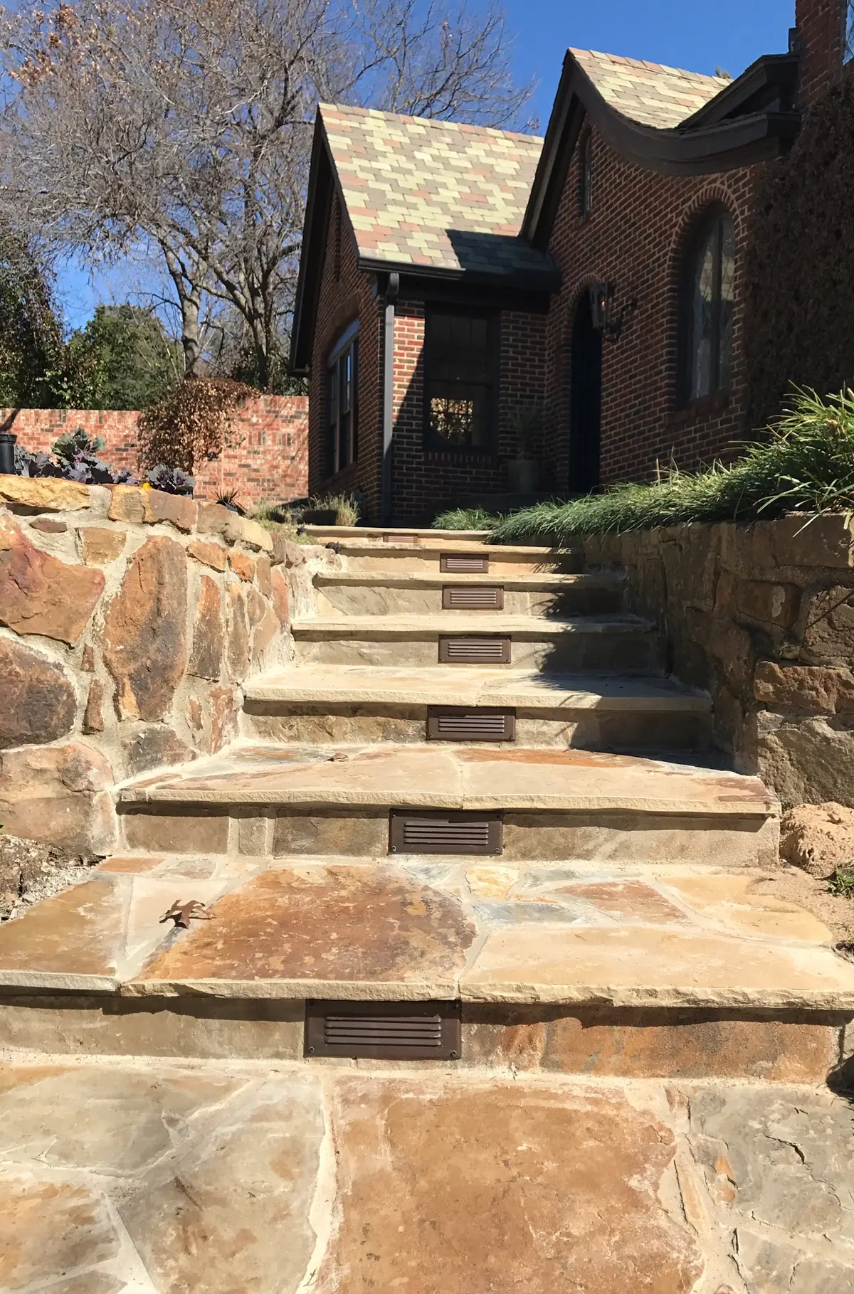 Flagstone steps with inset step lights between natural stone walls, leading up to a brick home
