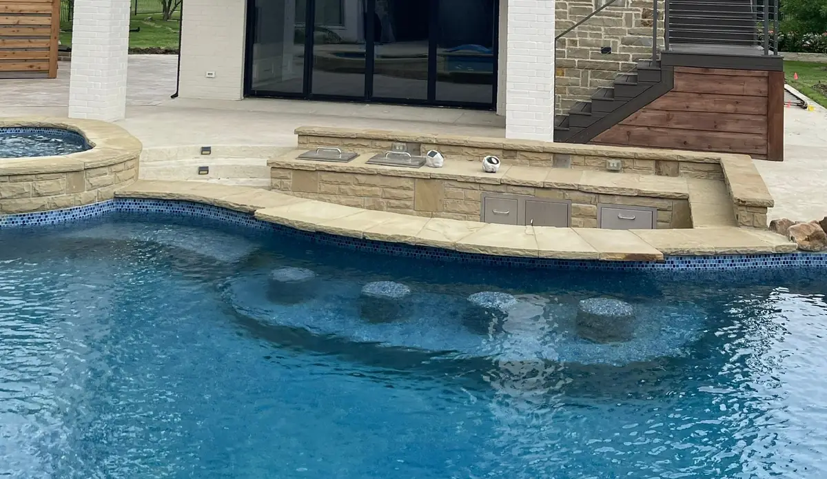 Stone pool coping and a stone grill bar beside swim-up seats in a blue pool