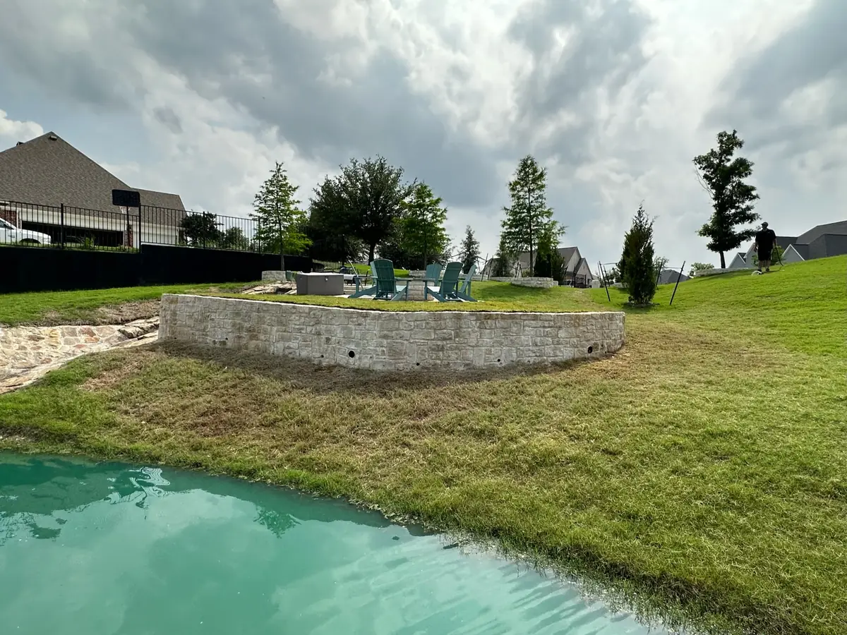 Curved stone retaining wall below a backyard patio at the edge of a pond